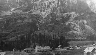 Photo of the town of Gothic, Colorado, dating from the 1880s. Buildings stand at the foot of a rocky mountain which has been dusted with snow. Most of the buildings are small one-story cabins, and one building in the center of the photo is a large white two-story building.
