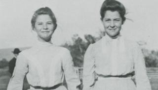 Photo of two young women standing side by side, outdoors. They are both wearing light colored, high neckline, long sleeved blouses with skirts, and they both wear their hair pulled up into buns on their heads. They are smiling at the camera.