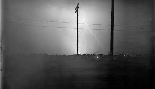 Photo of a large lightening bolt coming out of the cloud in the middle of the sky, and several fingers of lightening contact the ground. There are two very tall power line poles in the foreground. The bright lightening illuminates the darkness in this very dark photograph.