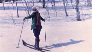 A woman stands on two skis in a snowy landscape. She is wearing a jacket, scarf, and hat, and looks over her shoulder at the camera.
