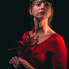 Photo of Amber Blais. She is illuminated against a black background, facing toward the spotlight. She wears a red leotard top and goggles on the top of her head, and she holds a bunch of long colorful feathers in her clasped hands. 