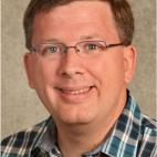 Photo of a man with short red hair that is parted on his left side, and he is wearing eyeglasses.  He wears a collared button-up shirt that is turquoise, navy, and gray plaid.