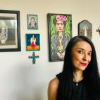 Photo of a woman standing in front of a white wall on which hangs several brightly colored images of the Virgin Mary, artist Frida Khalo, an ornate turquoise cross, another image of the Virgin Mary in a bright turquoise frame, and a macrame wall hanging made of crimson red yarns. The woman is dressed in a black dress and has long black hair with subtle stipes of gray on the top of her head.