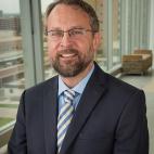 Photo of Doctor Mark Earnest. He is wearing a solid colored navy blue suit with a light blue dress shirt and a blue, yellow, and white gingham-patterned necktie. He has short sandy brown hair, with a beard and moustache of the same color. He wears eyeglasses and is smiling. He is standing in front of the large glass windows of a high-rise professional building, and other buildings are visible through the window.
