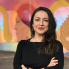 Photo of a woman with long dark hair, standing with her arms crossed in front of her.  She is wearing a black long-sleeved shirt and sleek gold drop earrings. She stands in a parking lot, with a very colorful mural painted on the wall in the distance behind her.