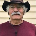 Photo of Robert Moll. He is standing in front of a building with yellow painted siding. Robert is wearing a burgundy colored t-shirt, with an outback-style dark leather hat. The had has a curled up brim on each side of his head, and there are cords from each side of the hat so that it can be tightened up under his chin. He has white curly short hair and a white moustache, and wears gold metal framed eyeglasses.
