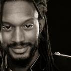 Photo of African American poet Stephen Brackett. Photo is a close up of his face and part of his left shoulder.  He has long hair in dreadlocks, and a shorter beard and moustache, and a warm, friendly expression on his face.