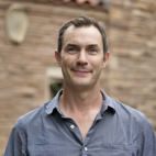 Photo of Thomas Andrews. This image shows a man wearing a blue button-up shirt with a relaxed collar, and the top two buttons are unbuttoned. He has short brown hair and wears no eyewear or facial hair.  He is standing outdoors, where a blurry background indicates a reddish brick building with ceramic tiles on the roof.
