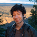 Photo of a young man standing outdoors, a vast sunny landscape framed on each side by tall pine trees behind him.  He has shorter, black wavy hair and is wearing a maroon colored t-shirt and a hooded windbreaker jacket. He is looking at the camera and smiling.