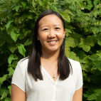Photo of Glenys Echavarri. She is standing outdoors, in front of a large green-leafed plant which is the entire background behind her. She is smiling, with her hands down at her sides. She is wearing a white blouse and a silver pendant. She has long straight dark colored hair that is parted on one side and hangs below her shoulders. 