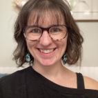 Portrait photograph of Julie Peterson. She wears glasses, dangling earrings, has dark hair, white skin, and is wearing a black top. 