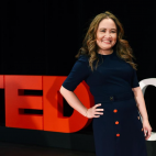 Photo of Nga Vương-Sandoval standing on a stage in front of a black background, and the large red letters that spell "TED" next to white letters "Ch" beginning the words "Che