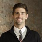 Photo of Sam Bock. He is wearing a dark suit jacket and a white dress shirt with a necktie. He has short dark hair and is smiling.