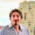Photo of a man with short but wavy, dark brown hair, moustache, and closely trimmed facial hair on his chin. He is wearing a black and white patterned button up shirt, with the first two buttons undone and the collar open casually. He is smiling. Behind him in the distance a couple of high rise buildings are visible. It is a bright day, and the sky is partly blue with some clouds.
