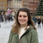 Photo of a woman named Allie Joyner. She is standing in a well-populated area, as lots of people are visible some distance behind her. It is chilly weather, as she and most others are wearing coats, some seem to be wearing a hat as well. This is an outdoor photo, with a large pine tree and multi-story buildings in the background. Allie has long dark hair with highlights at the ends. She is smiling. 