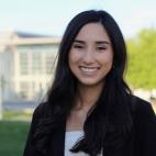 Photo of Raena Vigil, standing outdoors. Grass, trees, and a large building are in the distance behind.