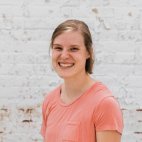 Photo of a young woman with short, straight blonde hair that is parted on the left side and brushed away from her face. She is smiling for the camera. She wears a peach colored V-neckline t-shirt with a small pocket to the left, and no jewelry or eyewear.