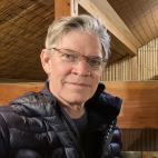 Photo of a middle-aged Caucasian man wearing eyeglasses. He has on a black shirt and a black puffy jacket. He has short-trimmed salt-and-pepper colored hair.