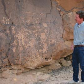 A tour member stands and admires a rock wall covered in large petroglyphs.