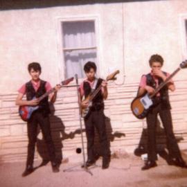 3 young men post in matching pink shirts and black vests. They each hold an electric guitar and pose for their performance.