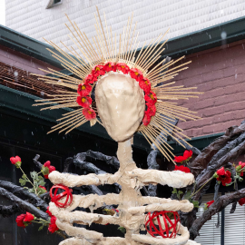 Artist Ariana “Nani” Gonzales next to her sculpture, Genesis. It shows a paper mache skeletal figure with a golden halo surrounded by roses, and dark branches.