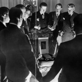 A group of men stand in a circle holding hands. There's a ghostly light shining on their faces from below.