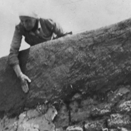 A woman leans over a tall edge of an adobe wall and applies a layer of mud.