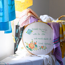 Drapes of colorful fabric fill the foreground as natural light focuses on an embroidery loop filled with flowers and the words: Thread my needle with its silk and men stacks of tired clothes.