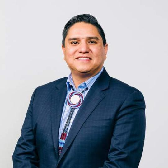Portrait photograph of Ernest House, Jr. He wears a beaded bolo tie, button down collared shirt, and a dark navy or black jacket.