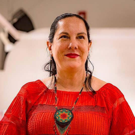 Photo of Adrianna Abarca, standing in front of a white background. She has long black hair that is pulled tightly to the back of her head, with dark eyes and red lips, she is smiling. She is wearing a bright red blouse and a beaded medallion necklace.