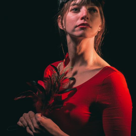 Photo of Amber Blais. She is illuminated against a black background, facing toward the spotlight. She wears a red leotard top and goggles on the top of her head, and she holds a bunch of long colorful feathers in her clasped hands. 