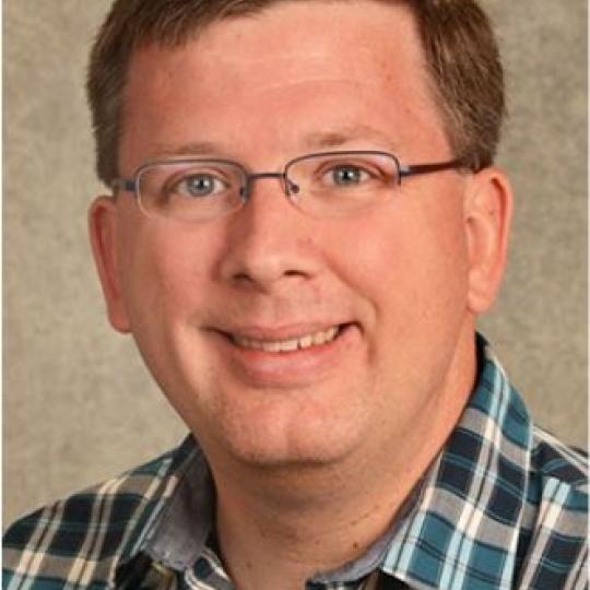 Photo of a man with short red hair that is parted on his left side, and he is wearing eyeglasses.  He wears a collared button-up shirt that is turquoise, navy, and gray plaid.