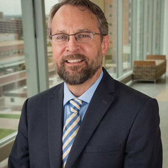 Photo of Doctor Mark Earnest. He is wearing a solid colored navy blue suit with a light blue dress shirt and a blue, yellow, and white gingham-patterned necktie. He has short sandy brown hair, with a beard and moustache of the same color. He wears eyeglasses and is smiling. He is standing in front of the large glass windows of a high-rise professional building, and other buildings are visible through the window.