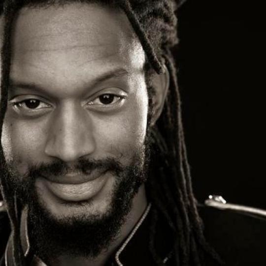 Photo of African American poet Stephen Brackett. Photo is a close up of his face and part of his left shoulder.  He has long hair in dreadlocks, and a shorter beard and moustache, and a warm, friendly expression on his face.