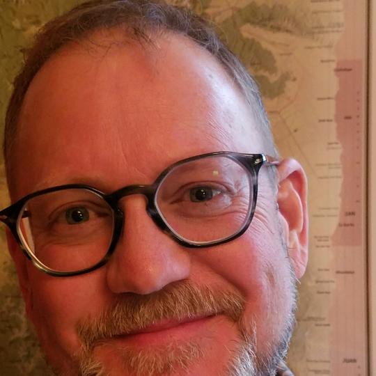 Photo of Steve Grinstead, author and managing editor of The Colorado Magazine. This is a close up photo of Steve. He has short, light brown hair, beard, and mustache. He is wearing a blue plaid collared shirt, gray tortoise shell glasses, and is smiling kindly. 