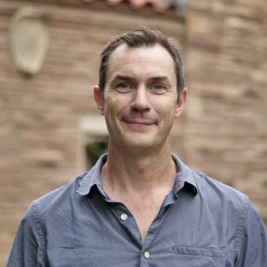 Photo of Thomas Andrews. This image shows a man wearing a blue button-up shirt with a relaxed collar, and the top two buttons are unbuttoned. He has short brown hair and wears no eyewear or facial hair.  He is standing outdoors, where a blurry background indicates a reddish brick building with ceramic tiles on the roof.