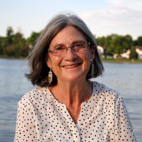 Photo of author Susan Casey. She is outdoors, and a lake with distant shore full of green trees is behind her. She has shoulder-length salt and pepper colored straight hair that is parted in the middle. She wears eyeglasses, and has long dangling earrings. She is smiling. Her blouse is white with small dark polka dots.