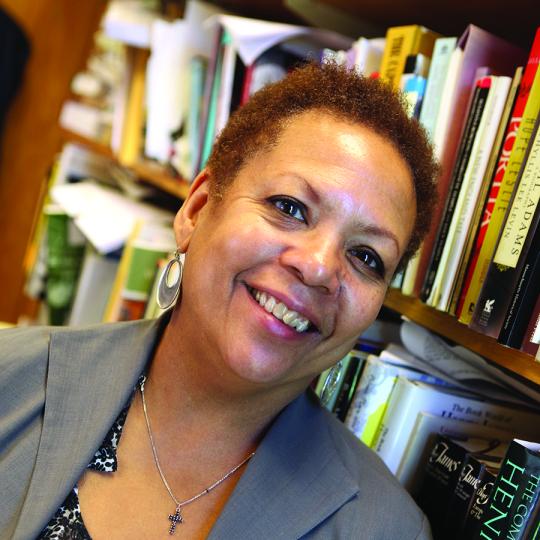 Photo of Claire Oberon Garcia, smiling at the camera and standing in front of book shelves full of books.  She has short curly reddish hair and is wearing silver dangling earrings and a silver chain with a small pendant. Her scoop neck blouse is decorated with flowers of varying shades of gray, and she has a gray jacket on over the top of her blouse.