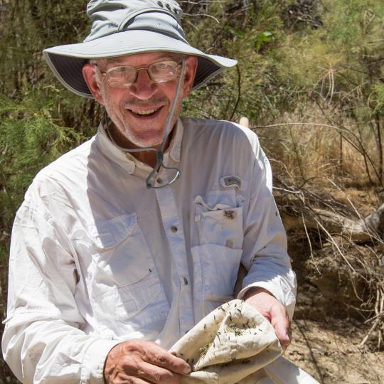 Photo of a man wearing a white long sleeve cotton button-up shirt, with lots of pockets on the front. He is smiling, and wearing a field hat with a wide brim and a cord that goes under his chin to secure the hat. He is holding a cloth that has a lot of small popcorn kernel sized bugs crawling around on it. He is outdoors near grasses and shrubs, on a sunny day.