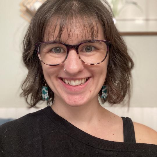 Portrait photograph of Julie Peterson. She wears glasses, dangling earrings, has dark hair, white skin, and is wearing a black top. 