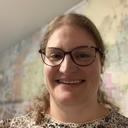 Photo of a woman with long blonde curly hair that is pulled away from her face. She is smiling and standing in a room with floral wallpaper on the wall behind her. She is wearing a black and white printed blouse and dark-rimmed eyewear.