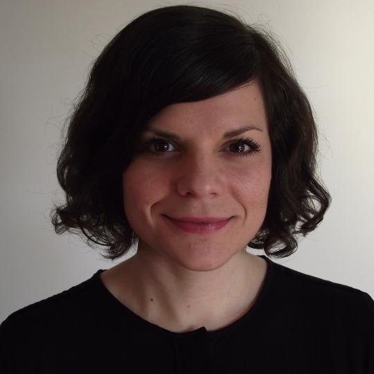 Photo of a woman with medium length, lightly curly black hair that is parted on the left side, and she is wearing a black scoop neck blouse. She is standing before a white background, that because of the lighting, looks black-to-gray on the left side behind her, and looks light-gray-to-white behind her on the right. She is smiling.