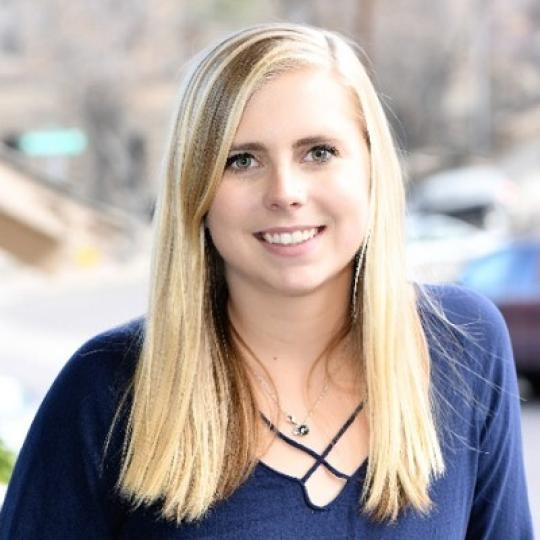 Photo of Natalie Gill. She wears a navy blue long sleeve blouse with a scoop neckline. She has long straight blonde hair that is parted on the left side of her head, and she has long bangs off to the right of her face. She is smiling.