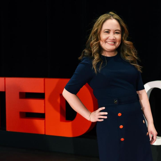 Photo of Nga Vương-Sandoval standing on a stage in front of a black background, and the large red letters that spell "TED" next to white letters "Ch" beginning the words "Che