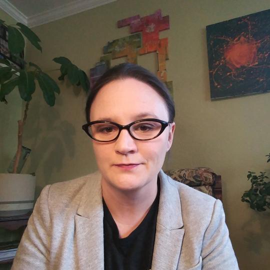 Photo of a woman sitting in a chair. She is wearing a black shirt with a gray jacket. She is not wearing any visible jewelry, but wears fashionable black cateye eyeglasses, and has her hair pulled back away from her face. Behind her are two sizeable green leafed plants, and on the wall hang two pieces of artwork that are brightly colored.