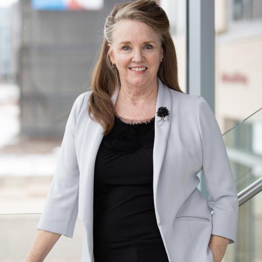 Photo of Peggy O'Neill-Jones. She is standing against a railing upon which her right hand is resting. Her left hand is in her left pocket. She is wearing a black blouse with black pants, and a light gray blazer that has a black rose brooch on the left lapel. She has long shoulder-length blonde hair that is parted on the right and is swept back away from her face. She is smiling.