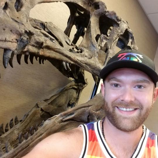 Photo of a man who appears to be taking a selfie in front of a T-Rex skeleton head. The man is smiling. He is wearing a black baseball cap with a colorful logo on the front, and a brightly-striped tank top, as well as a necklace of black beads. He has a light brownish, close-shaved beard.