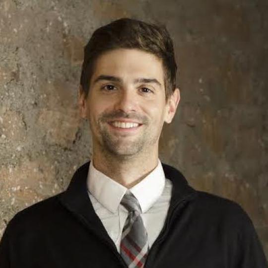 Photo of Sam Bock. He is wearing a dark suit jacket and a white dress shirt with a necktie. He has short dark hair and is smiling.