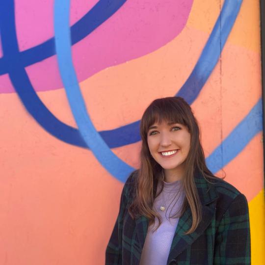 Photo of a young woman with long dark hair and bangs. She is smiling broadly while standing in front of a portion of very colorfully painted outdoor mural. The wall is orange and yellow, with a pink post and blue circles. She is wearing a purple turtle neck shirt, with a dark blue and green plaid coat that is unbuttoned in front, and a silver and pearl pendant around her neck.