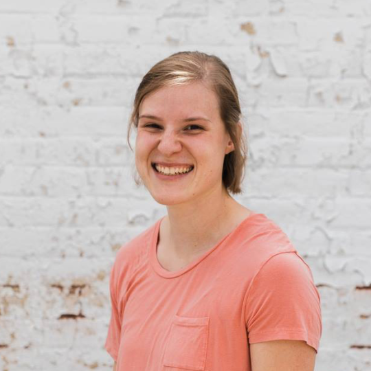Photo of a young woman with short, straight blonde hair that is parted on the left side and brushed away from her face. She is smiling for the camera. She wears a peach colored V-neckline t-shirt with a small pocket to the left, and no jewelry or eyewear.